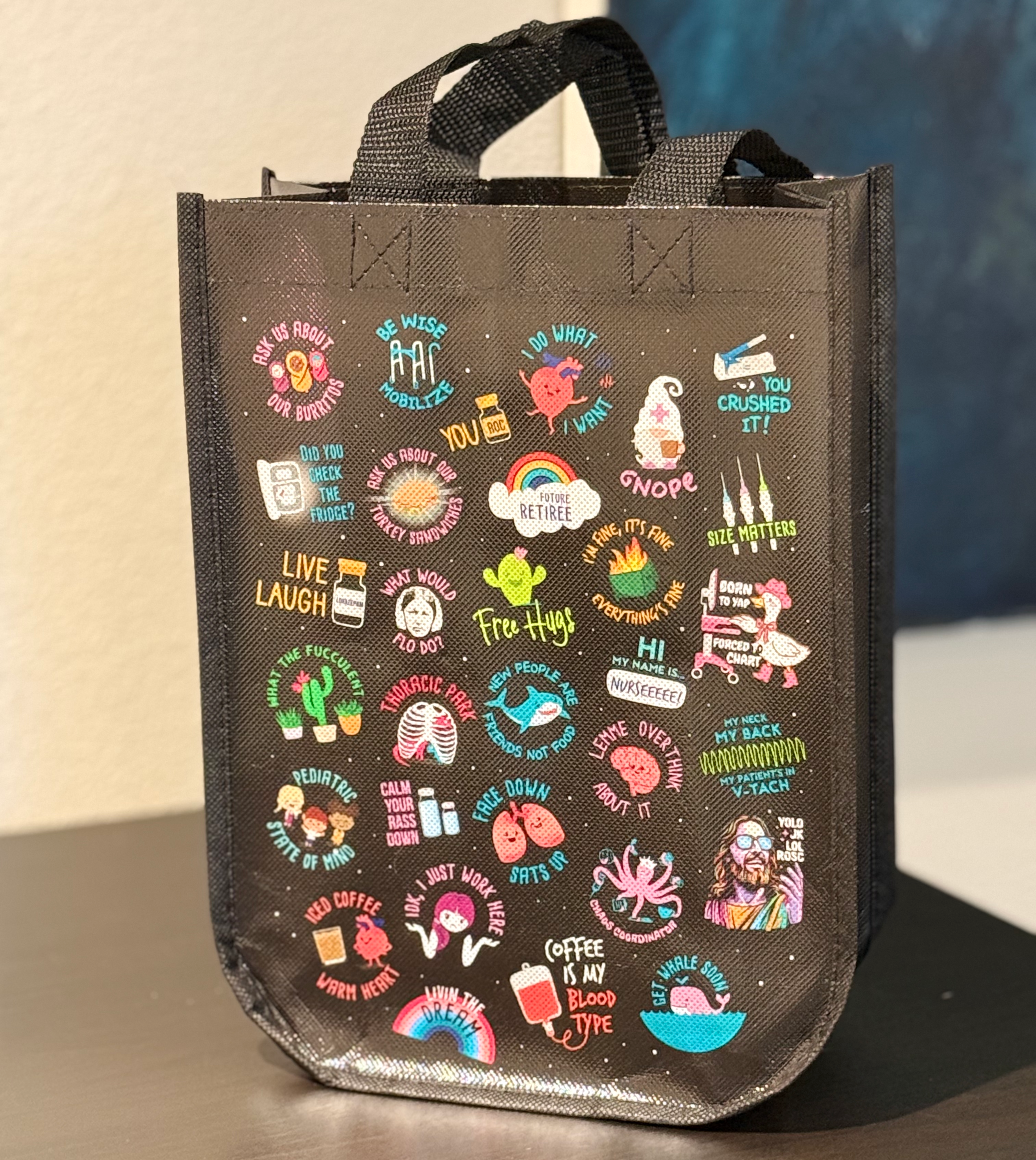 A black tote bag with a variety of colorful stickers and illustrations on it, including phrases like "Live Laugh", "Free Hugs", "Size Matters", and "I Just Work".